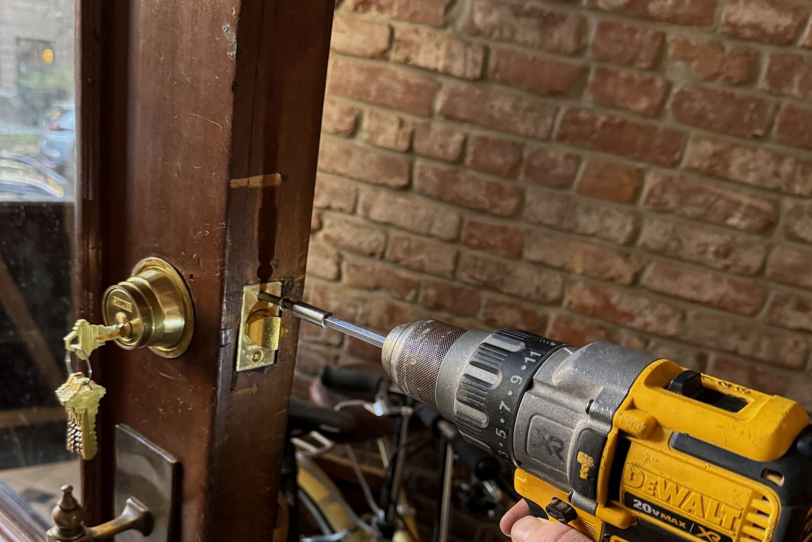 Cordless drill driving a screw into the latch plate of a wood door with a brass deadbolt and keys in the lock