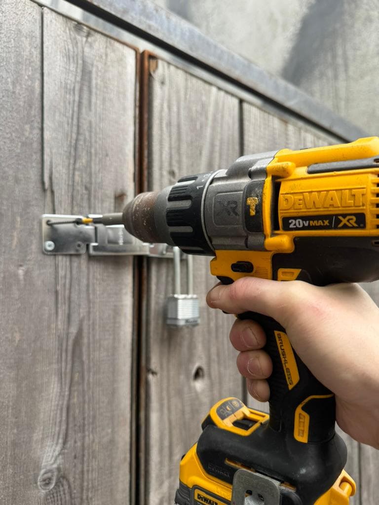 Cordless drill fitting a hasp on a weathered wooden gate with a padlock hanging below