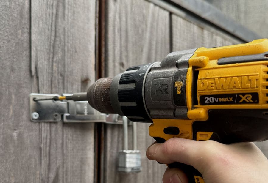 Cordless drill driving a screw into a metal hasp on a weathered wooden gate, padlock below
