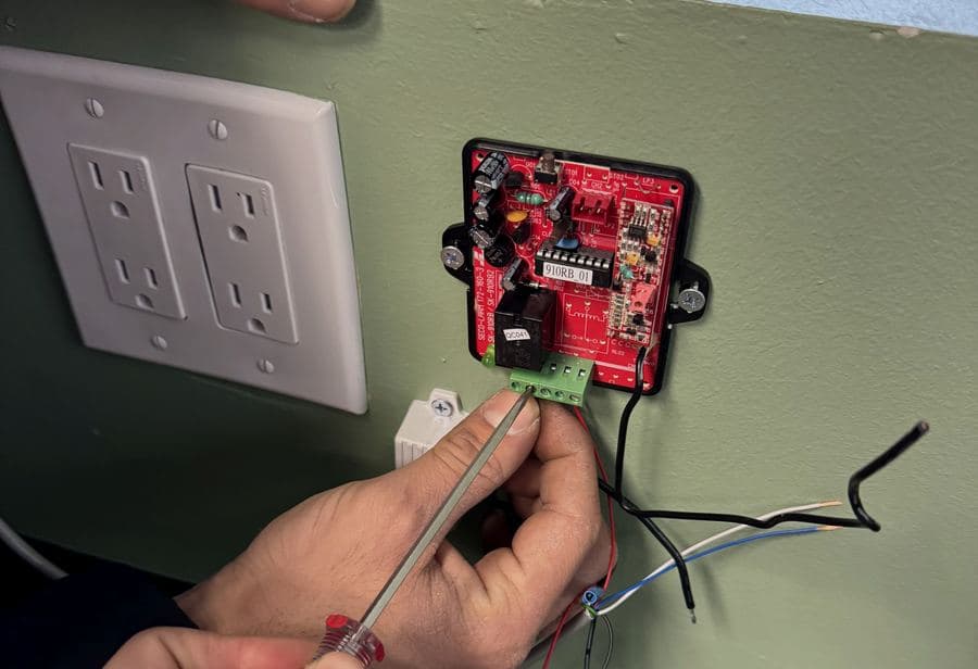 Red relay circuit board with a green terminal block being wired with a screwdriver on a wall