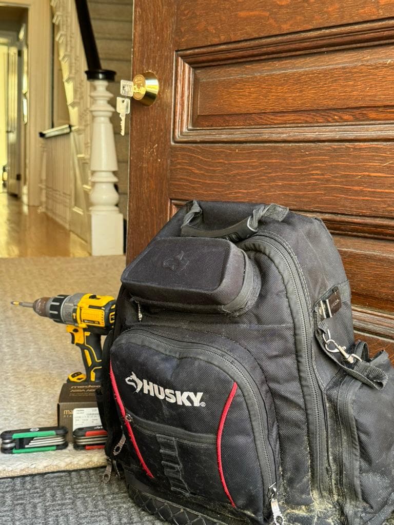 Tool backpack and cordless drill on the floor in front of a dark wood door with a brass deadbolt and keys in it